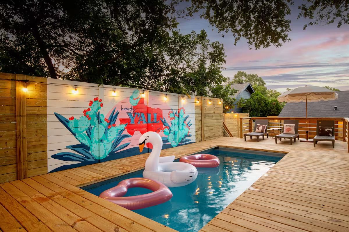 Sunset backyard wooden deck with a rectangular plunge pool, a large inflatable swan and two pink donut floats, string lights and a colorful cactus mural on the fence reading Y'ALL, plus lounge chairs and an umbrella