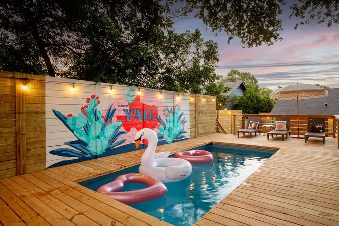 Sunset backyard wooden deck with a rectangular plunge pool, a large inflatable swan and two pink donut floats, string lights and a colorful cactus mural on the fence reading Y'ALL, plus lounge chairs and an umbrella
