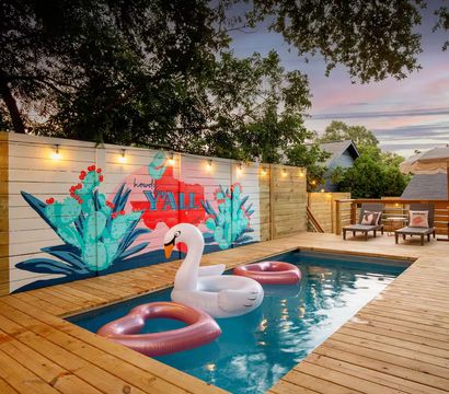 Sunset backyard wooden deck with a rectangular plunge pool, a large inflatable swan and two pink donut floats, string lights and a colorful cactus mural on the fence reading Y'ALL, plus lounge chairs and an umbrella