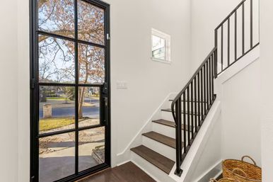 Welcoming modern foyer with a black grid glass front door, dark wood staircase and black metal railing, white walls and woven baskets