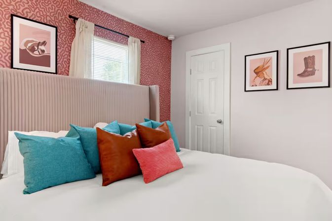 Stylish cozy bedroom interior with white bedding, ribbed upholstered headboard, teal and rust decorative pillows, pink patterned accent wall, small curtained window and framed western artwork
