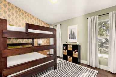 Bright kids' bedroom with dark wood bunk bed, cactus-print accent wallpaper, sage-green walls, striped rug, black cube storage with woven baskets, and sunlight streaming through a curtained window.