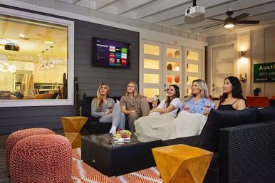 Five friends relaxing on a covered Austin patio living room, cozy sectional with blankets and poufs, holding wine and watching a wall-mounted TV/streaming projector with snacks on the glass coffee table — modern indoor-outdoor home theater vibe.
