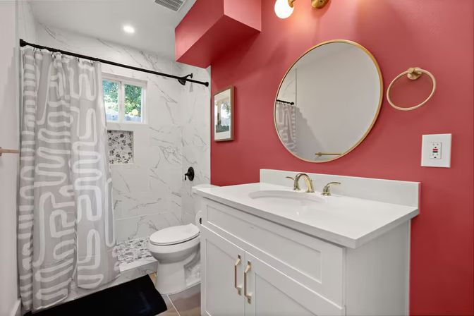 Modern residential bathroom with coral-red accent wall, white vanity and gold fixtures, round mirror, marble-tiled walk-in shower with patterned gray curtain and small window
