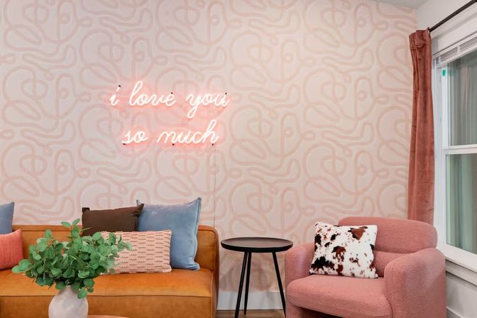 Cozy modern living room with a tan leather sofa, pastel abstract wallpaper and pink neon sign reading “i love you so much,” pink boucle armchair with cowhide pillow, black side table, and vase of eucalyptus by a window.