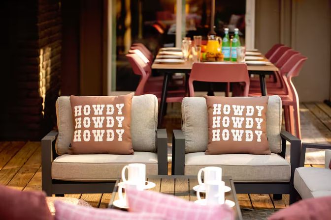 Outdoor wooden deck patio with two cushioned lounge chairs featuring brown pillows printed HOWDY HOWDY HOWDY, a coffee table with white mugs, and a dining table with pink chairs and bottled drinks in the background.