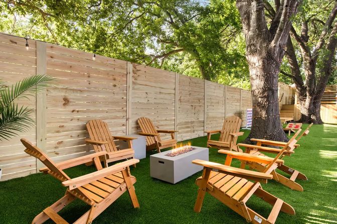 Shaded backyard lounge with wooden Adirondack chairs arranged around a rectangular fire pit on lush green turf, tall wooden privacy fence, large oak trees and string lights.