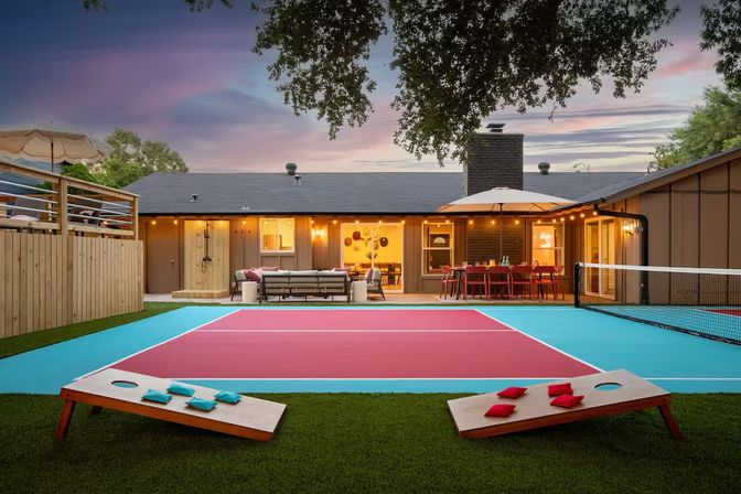 Vibrant backyard at dusk with a red-and-turquoise painted sport court (pickleball-style), cornhole boards on artificial turf, cozy outdoor sofa, umbrella-shaded dining table, twinkling string lights and a single-story patio house