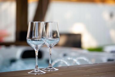 Two empty wine glasses on a wooden ledge beside a bubbling outdoor hot tub on a sunlit backyard patio
