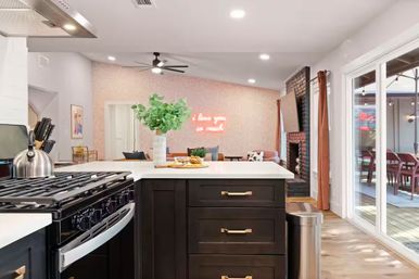Bright modern open-concept kitchen and living room with a dark island and white countertop, stainless steel stove, vase of greenery, neon wall sign, fireplace and sliding glass doors to a patio deck.