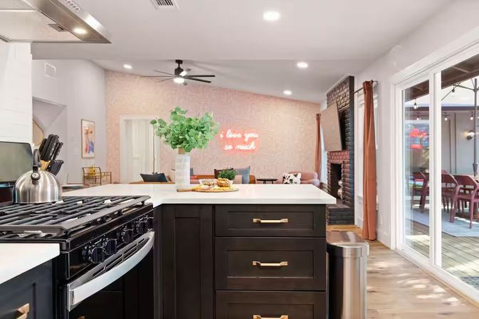 Bright modern open-concept kitchen and living room with a dark island and white countertop, stainless steel stove, vase of greenery, neon wall sign, fireplace and sliding glass doors to a patio deck.