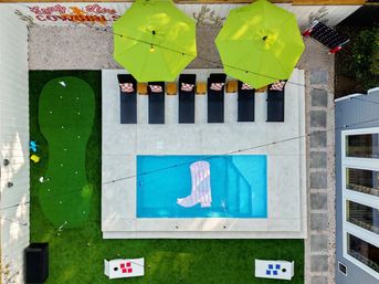 Aerial view of a residential backyard: small rectangular blue pool with a pink boot float, concrete deck lined with black loungers under lime-green umbrellas, adjacent putting green, cornhole boards and string lights.