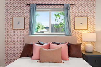 Sunlit cozy bedroom with beige upholstered headboard, pink and brown throw pillows, playful horseshoe wallpaper, blue curtains framing a window with a backyard view, framed art and bedside lamp.