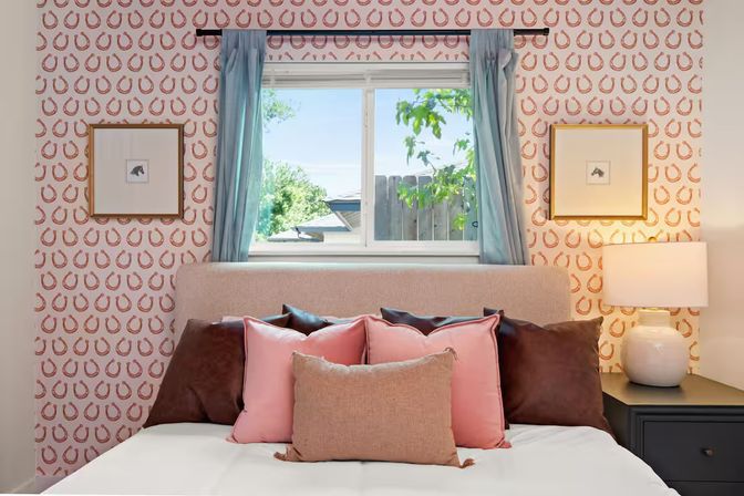 Sunlit cozy bedroom with beige upholstered headboard, pink and brown throw pillows, playful horseshoe wallpaper, blue curtains framing a window with a backyard view, framed art and bedside lamp.