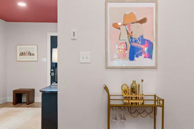 Modern home entryway decor: pink ceiling, abstract portrait above a gold bar cart with cocktail tools and hanging wine glasses, with a small bench and framed city print in the adjoining room.