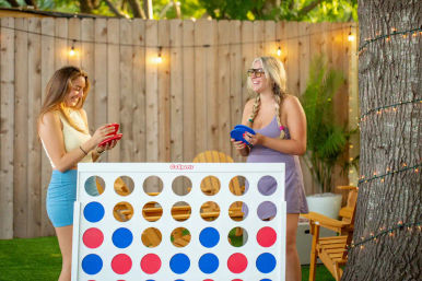 Two women laughing while playing a giant Connect Four lawn game on a grassy backyard patio, holding red and blue discs next to a wooden fence, string lights, Adirondack chairs and a tree wrapped in lights.