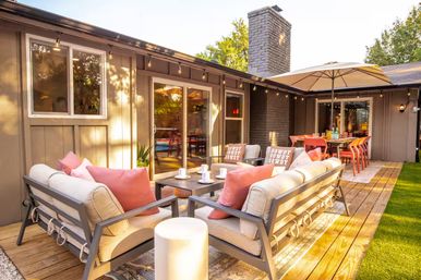 Sunlit suburban backyard deck with cushioned outdoor sofas and coral pillows around a coffee table, string lights overhead and umbrella‑shaded dining table by sliding glass doors.