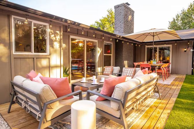 Sunlit suburban backyard deck with cushioned outdoor sofas and coral pillows around a coffee table, string lights overhead and umbrella‑shaded dining table by sliding glass doors.