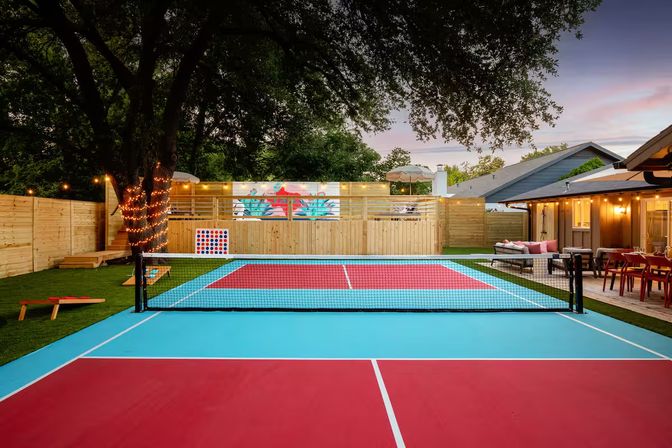 Vibrant red-and-blue backyard pickleball court at dusk, fenced yard with a string-lit oak tree, outdoor dining and lounge area, mural and cornhole game.