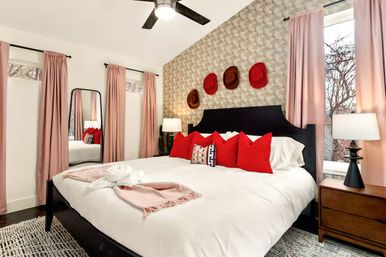 Cozy contemporary master bedroom with large white bed, black headboard, bright red throw pillows, patterned accent wall with mounted hats, pink floor-length curtains framing windows, ceiling fan, full-length mirror, and wooden nightstand with lamp.
