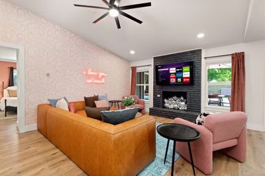 Bright modern living room with a tan leather sectional, pink upholstered chairs, patterned pink accent wall featuring a neon 'I love you so much' sign, black brick fireplace with wall-mounted TV and decorative metallic orbs, hardwood floors, ceiling fan, and windows opening to a backyard patio.