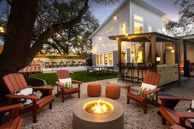 Cozy evening backyard patio at a modern farmhouse-style home featuring a glowing round fire pit surrounded by wooden Adirondack chairs, woven poufs, string lights draped from large oak trees, a pergola bar with stools, and a pool with lounge seating.