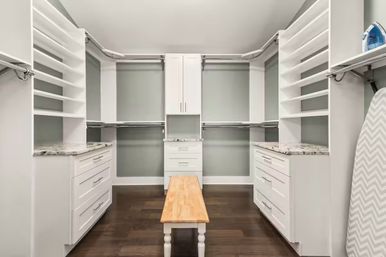 Spacious modern walk-in closet with custom white built-in drawers and open shelves, marble-look countertops, dual hanging rods, dark hardwood floor, wooden bench centered and ironing board at right.