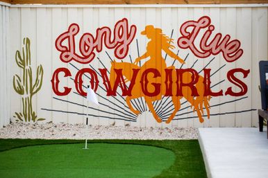 Backyard scene with a Western-themed mural on a white wooden fence reading 'Long Live Cowgirls', orange cowgirl-on-horse silhouette, cactus illustration, and a small green putting green with a flag in the foreground.