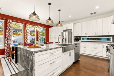Bright open-concept modern kitchen with white shaker cabinets, gray marble island with farmhouse sink, stainless steel appliances, three black pendant lights, red accent wall, dining area by large windows and hardwood floors.
