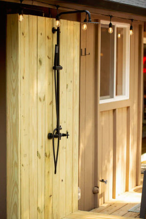Outdoor backyard shower with black matte rain showerhead and handheld mounted on a light wooden privacy panel, string bulb patio lights and adjacent deck siding.
