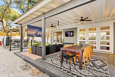 Covered backyard patio with long wooden dining table and bright orange chairs on a geometric rug, adjacent wicker lounge with wall-mounted TV and ceiling fans — modern outdoor entertaining and dining space.