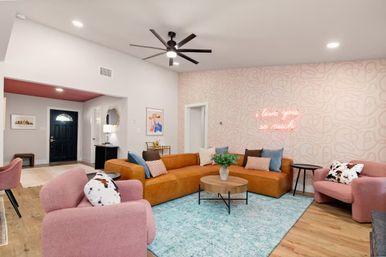 Stylish modern living room with a tan leather sectional, blush-pink armchairs, teal area rug and round wood coffee table, patterned pink accent wall with neon 'i love you so much' sign, wood floors and black ceiling fan.