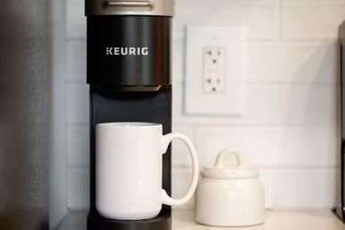 Single-serve coffee maker on a kitchen counter with a white ceramic mug under the spout, a small sugar jar, and white subway tile backsplash