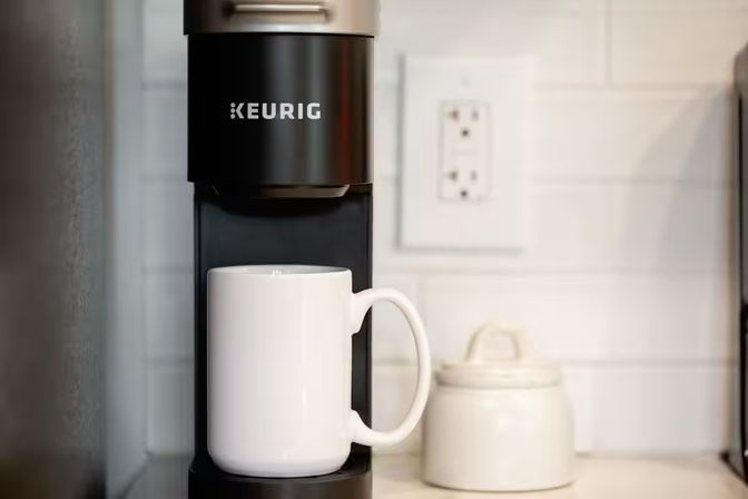 Single-serve coffee maker on a kitchen counter with a white ceramic mug under the spout, a small sugar jar, and white subway tile backsplash