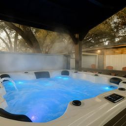 Steaming blue-lit outdoor hot tub under a wooden pergola at dusk, bubbling water with a waterfall spout and cozy backyard seating framed by trees.