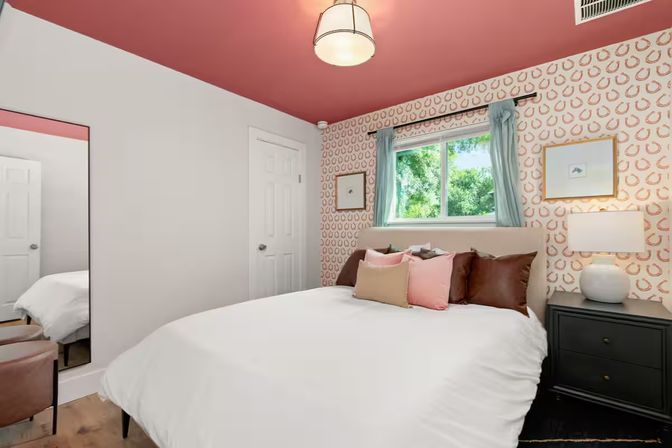 Cozy modern guest bedroom with white duvet, pink and tan throw pillows, coral-patterned wallpaper and matching coral ceiling, window overlooking a leafy yard, and a black nightstand with a white lamp.