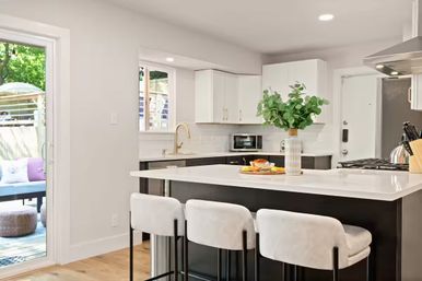Bright modern open-concept kitchen with white cabinets and black island topped with white quartz, three white bar stools, brass faucet, vase of green eucalyptus, stainless range, and sliding door opening to a leafy backyard patio.