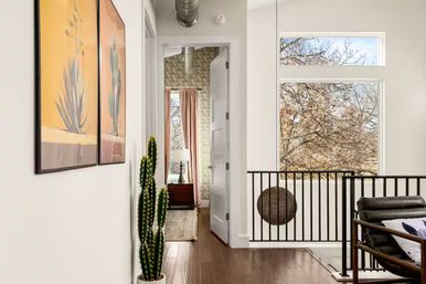 Bright modern upstairs landing in a contemporary home with hardwood floors, tall potted cactus, framed botanical prints, black metal railing, large windows overlooking fall trees, and a bedroom doorway with pink curtains and bedside lamp.