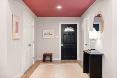 Modern home entryway/foyer with a black front door and arched window, blush-pink ceiling, light gray walls, wood floor, neutral rug, small bench, and a console table with lamp and mirror.