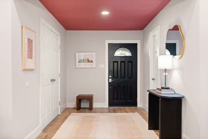 Modern home entryway/foyer with a black front door and arched window, blush-pink ceiling, light gray walls, wood floor, neutral rug, small bench, and a console table with lamp and mirror.