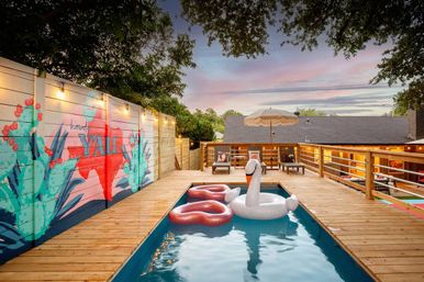Backyard wooden deck with a narrow plunge pool at sunset, inflatable swan and two heart-shaped floats, colorful cactus mural on the fence reading Howdy Y'ALL, string lights, lounge chairs and umbrella