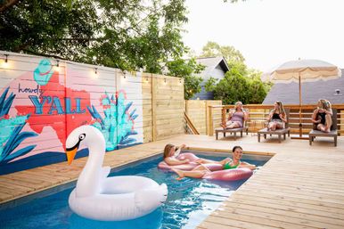 Sunny backyard wooden deck with a small rectangular pool, a giant white swan float, two people lounging on pink inflatables in the water, three people relaxing on lounge chairs under an umbrella, and a colorful fence mural reading 'howdy Y'ALL' with string lights.