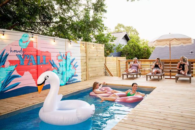 Sunny backyard wooden deck with a small rectangular pool, a giant white swan float, two people lounging on pink inflatables in the water, three people relaxing on lounge chairs under an umbrella, and a colorful fence mural reading 'howdy Y'ALL' with string lights.