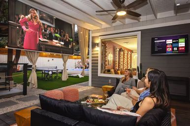 Cozy modern backyard patio entertainment area with covered lounge, large outdoor projector screen showing a courtroom scene, three women on a black sectional enjoying wine and charcuterie, ping-pong table and wall-mounted smart TV for an alfresco movie night.