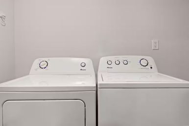 Side-by-side white washing machine and dryer in a clean home laundry room with neutral gray walls and a visible electrical outlet
