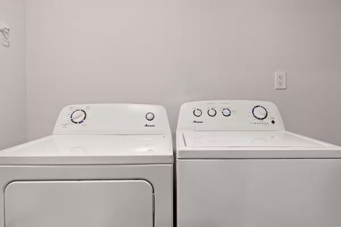 Side-by-side white washing machine and dryer in a clean home laundry room with neutral gray walls and a visible electrical outlet