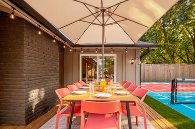 Sunny backyard patio with a wooden dining table set for eight, coral chairs and a large umbrella, string lights and sliding glass door, beside a vibrant blue-and-red sports court and fenced yard.