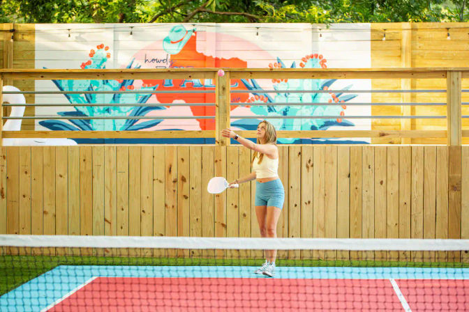Player serving a pickleball on a bright outdoor court with red-and-blue surface, wooden fence, leafy trees and a colorful tropical mural