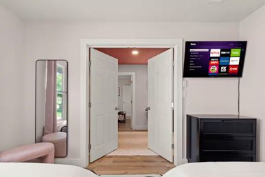 Bright modern bedroom with double white doors opening to a hallway, wall-mounted smart TV showing streaming apps, black dresser, tall mirror and pink ottoman.