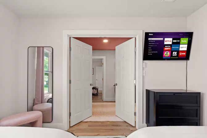 Bright modern bedroom with double white doors opening to a hallway, wall-mounted smart TV showing streaming apps, black dresser, tall mirror and pink ottoman.